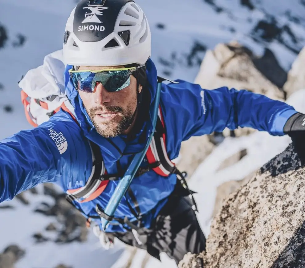 A climber wearing a white helmet with "SIMOND" branding and a blue jacket with "The North Face" logo ascends a rocky, snow-covered mountain, gripping the terrain with gloved hands and supported by climbing gear.