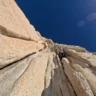 A rock climber ascends a steep granite cliff under a clear blue sky, with sunlight creating lens flares. The climber is secured with a red rope, navigating a narrow vertical crack in the rock face.