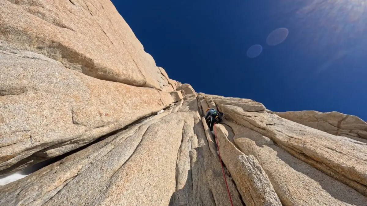 A rock climber ascends a steep granite cliff under a clear blue sky, with sunlight creating lens flares. The climber is secured with a red rope, navigating a narrow vertical crack in the rock face.