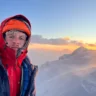 A person wearing a red and blue winter jacket and an orange helmet with frost on their face, standing on a snowy mountain ridge during a colorful sunrise or sunset with clouds and distant peaks in the background.