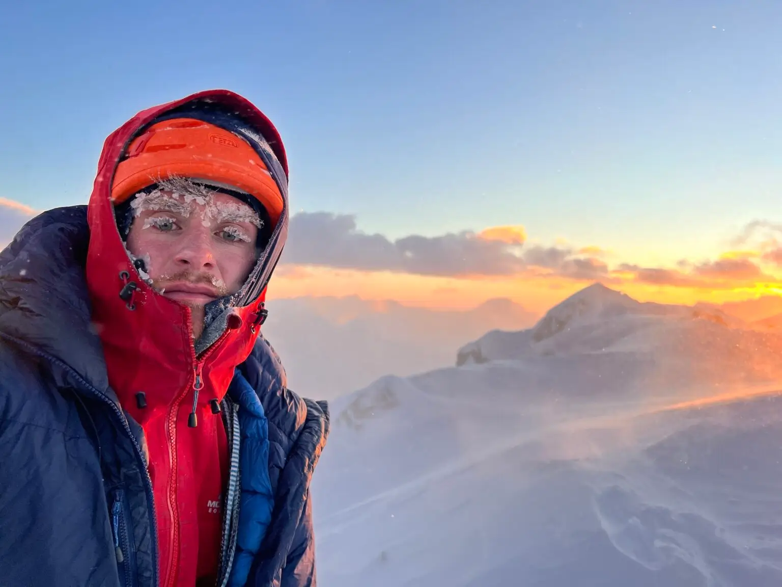 A person wearing a red and blue winter jacket and an orange helmet with frost on their face, standing on a snowy mountain ridge during a colorful sunrise or sunset with clouds and distant peaks in the background.