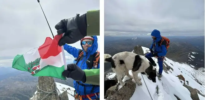 A person in a blue jacket and gloves holds a flag with green, white, and red vertical stripes featuring a coat of arms, standing on a snowy mountain peak with a dog nearby, surrounded by a vast mountainous landscape under a cloudy sky.