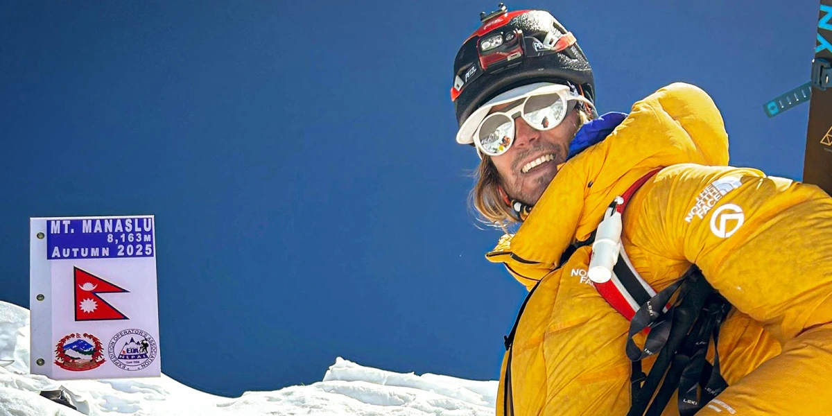 A person in a bright yellow mountaineering jacket and helmet stands on a snowy ridge with a clear blue sky in the background. To the left, a sign reads "MT. MANASLU 8,163m AUTUMN 2025" with the Nepalese flag and a logo of the Nepal Mountaineering Association. The person is equipped with climbing gear, including ropes and a harness.