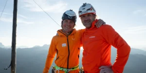 Two climbers wearing orange jackets and helmets stand with arms around each other, smiling, against a backdrop of mountains under a clear sky with a wooden pole and cables nearby.
