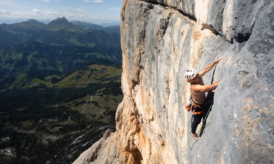 A shirtless climber in a helmet and harness ascends a steep rock face, with a scenic view of green hills and distant mountains under a partly cloudy sky.