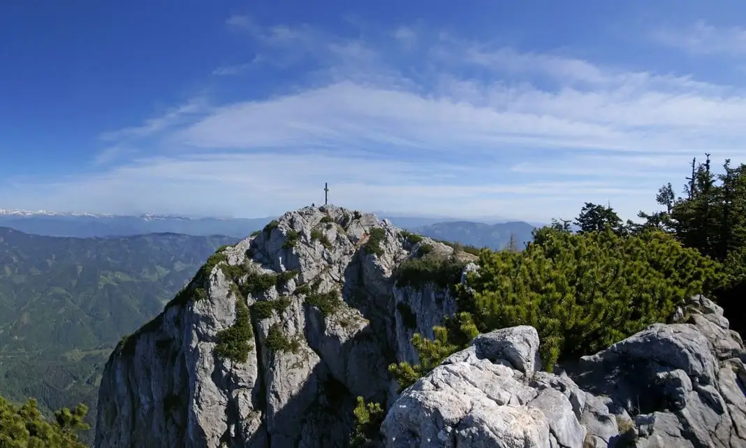 A rugged mountain peak with a cross at the summit, surrounded by rocky terrain and green shrubs. The background features a vast range of rolling hills and distant snow-capped mountains under a clear blue sky with wispy clouds.