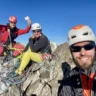 Three climbers in helmets and climbing gear pose for a selfie on a rocky mountain peak, with ropes and gear scattered around. One climber in a red jacket and red helmet raises a fist, another in a white helmet and yellow pants sits relaxed, and the third in an orange and white helmet smiles at the camera. In the background, a steep mountain face with a red-marked climbing route labeled "Мишрин (3) 5018 м/н.ч.м" and a glacier below are visible under a clear blue sky.