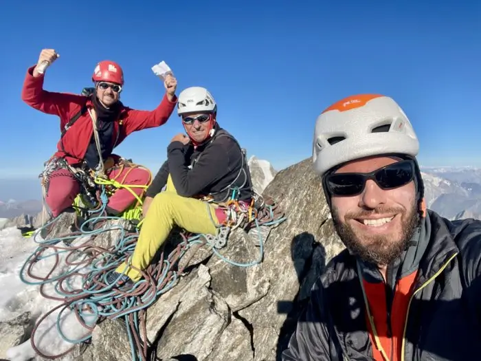 Three climbers in helmets and climbing gear pose for a selfie on a rocky mountain peak, with ropes and gear scattered around. One climber in a red jacket and red helmet raises a fist, another in a white helmet and yellow pants sits relaxed, and the third in an orange and white helmet smiles at the camera. In the background, a steep mountain face with a red-marked climbing route labeled "Мишрин (3) 5018 м/н.ч.м" and a glacier below are visible under a clear blue sky.