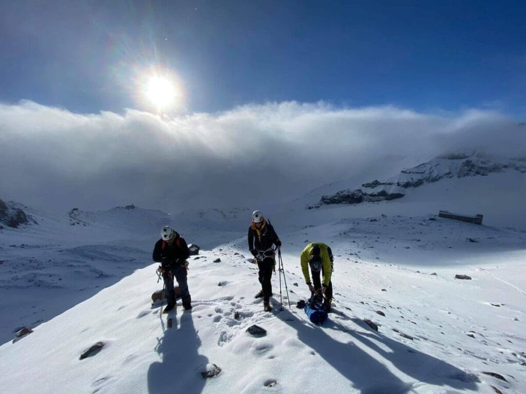 Three climbers with helmets and gear standing on a snowy ridge under a bright sun partially obscured by clouds, with one person adjusting a blue bag and mountains in the background.