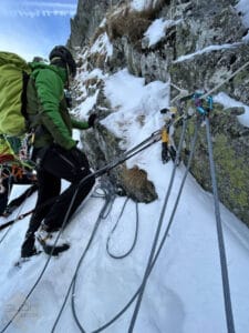 A climber in a green jacket and helmet ascending a snowy, rocky cliff, secured with multiple ropes and climbing gear attached to the rock face, under a clear blue sky.