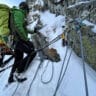 A climber in a green jacket and helmet ascending a snowy, rocky cliff, secured with multiple ropes and climbing gear attached to the rock face, under a clear blue sky.