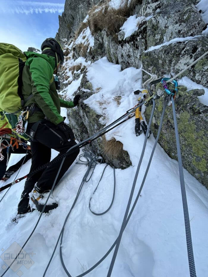 A climber in a green jacket and helmet ascending a snowy, rocky cliff, secured with multiple ropes and climbing gear attached to the rock face, under a clear blue sky.