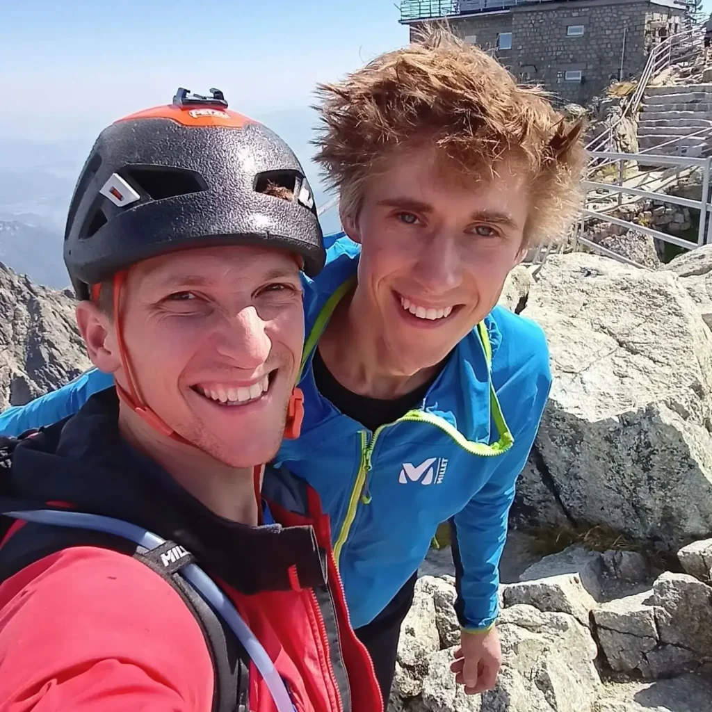Two people wearing climbing gear, including helmets and jackets, smiling for a selfie on a rocky mountain summit with a stone building and railing visible in the background under a clear blue sky.