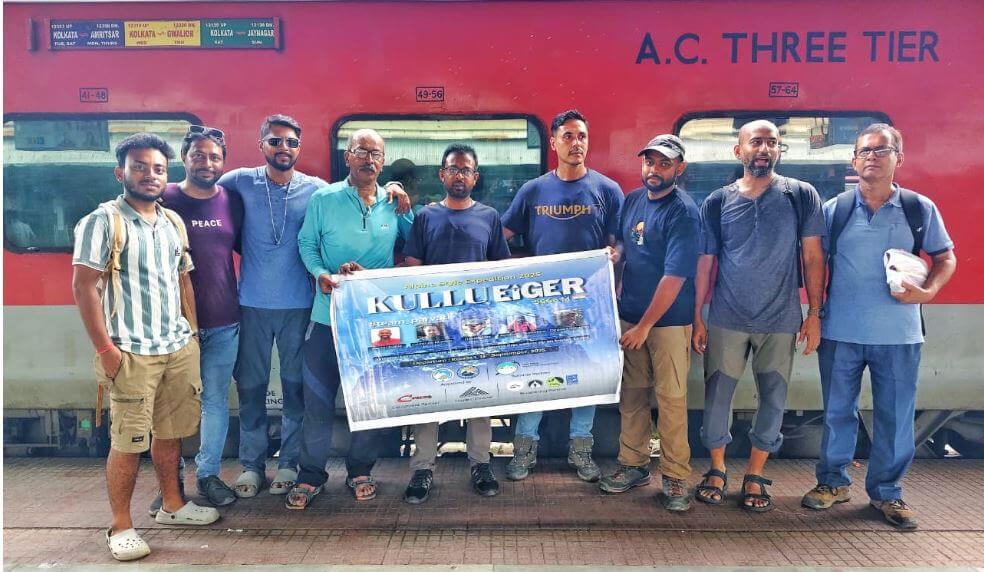 A group of eight people standing in front of a red AC Three Tier train, holding a banner that reads "KulluEiger" with images and text related to an expedition. The individuals are dressed in casual and outdoor clothing, some wearing hats and backpacks, suggesting a hiking or trekking group. The train has windows and a sign with destination and timing information.