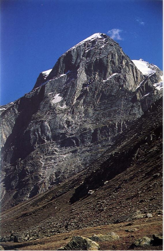 A towering, rugged mountain with a snow-capped peak rises against a clear blue sky. The steep, rocky slopes are partially covered with patches of snow, and the foreground features a barren, rocky terrain with scattered stones and sparse vegetation.