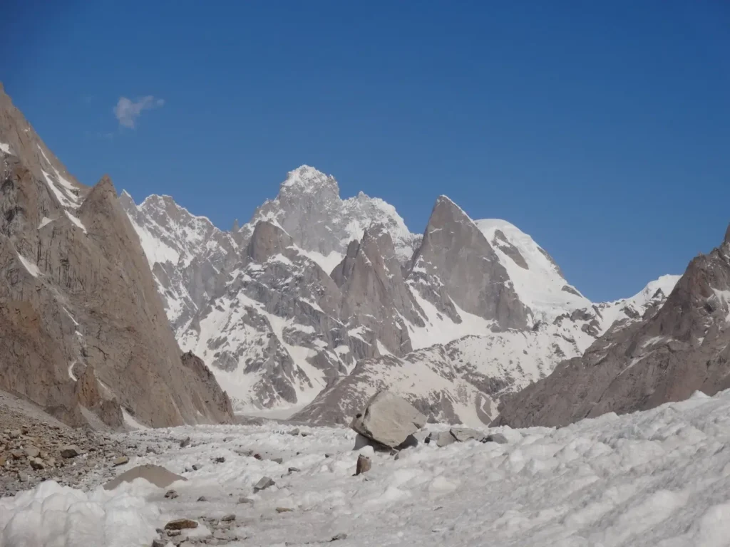 A scenic view of a snow-covered mountain range under a clear blue sky. The foreground features a glacier with scattered rocks, and the rugged peaks rise sharply in the distance.