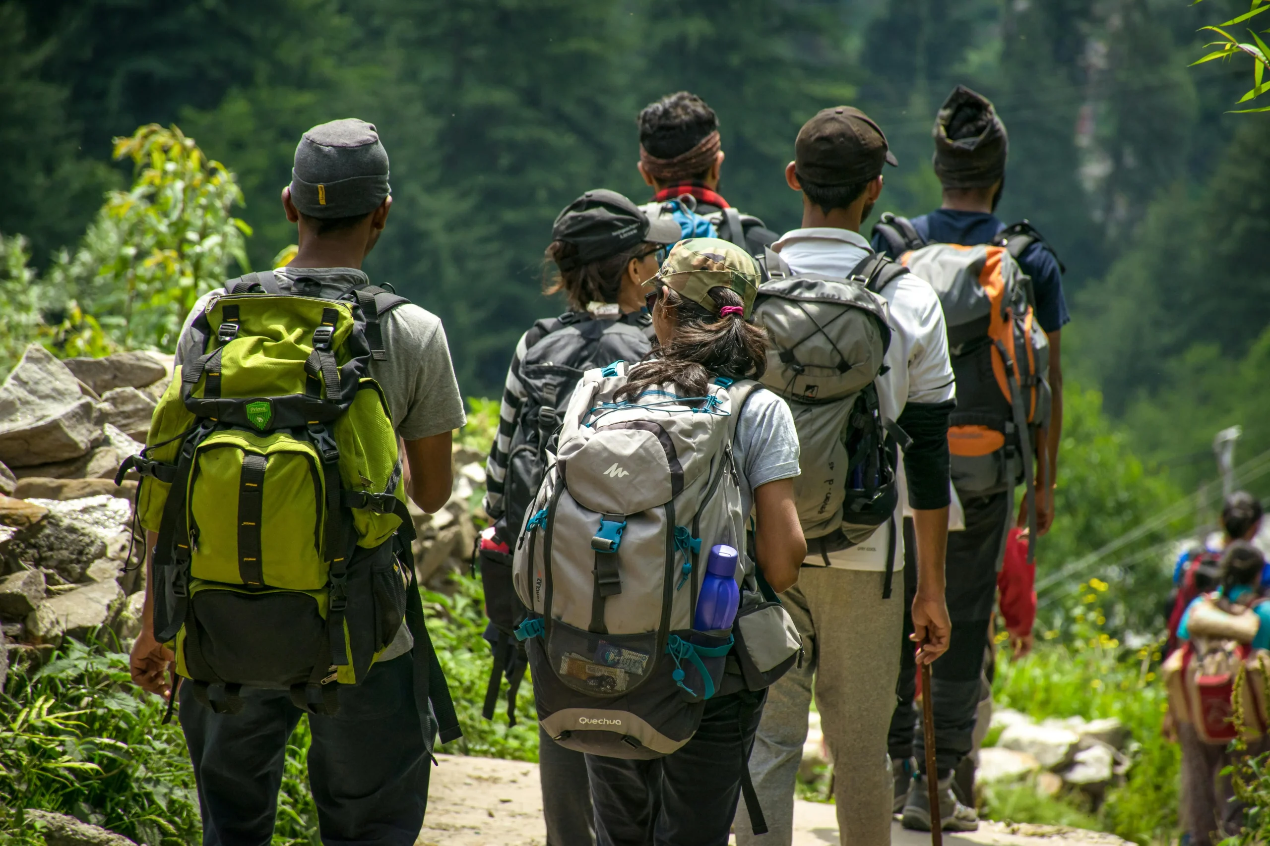 A group of hikers walking along a narrow trail in a lush green forest, each carrying large backpacks, with dense vegetation and rocky terrain on either side.