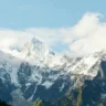 A stunning view of a snow-covered mountain range under a clear blue sky with scattered clouds, featuring rugged peaks and green valleys below.