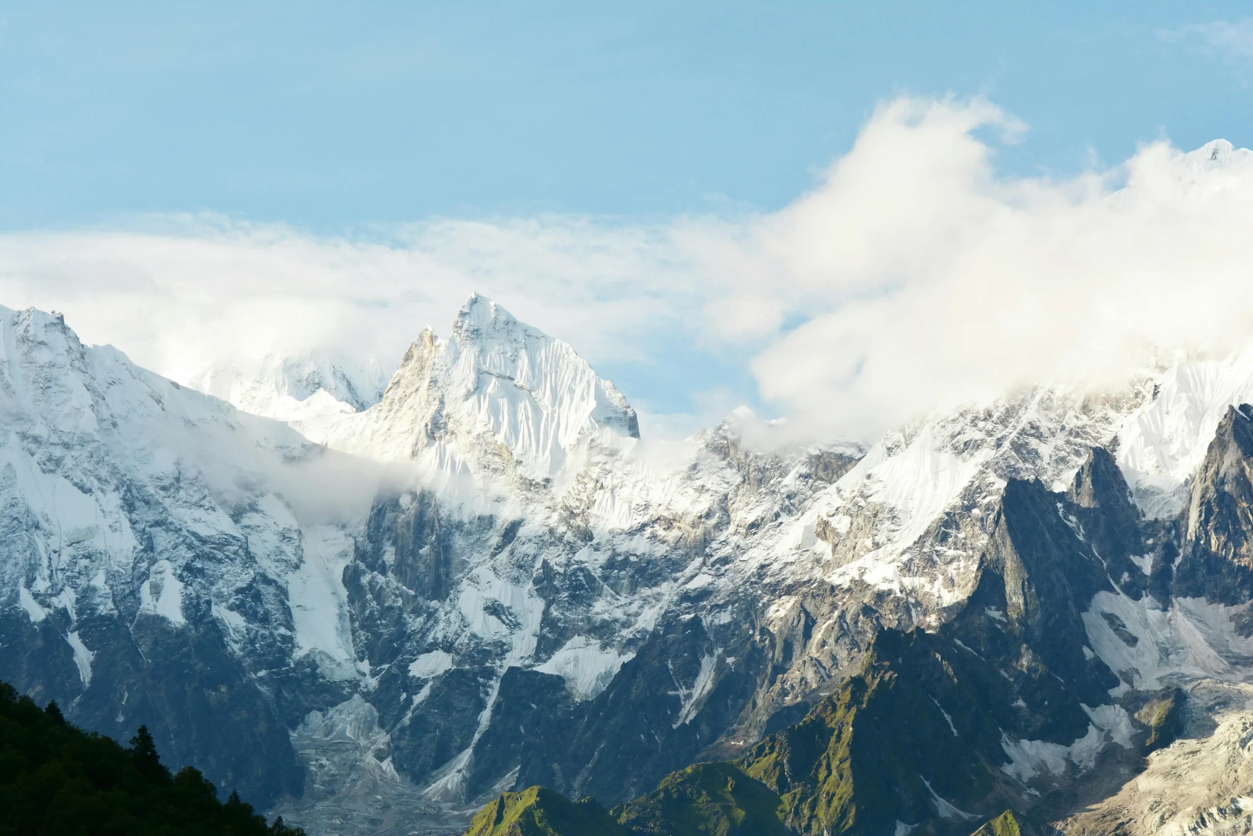 A stunning view of a snow-covered mountain range under a clear blue sky with scattered clouds, featuring rugged peaks and green valleys below.