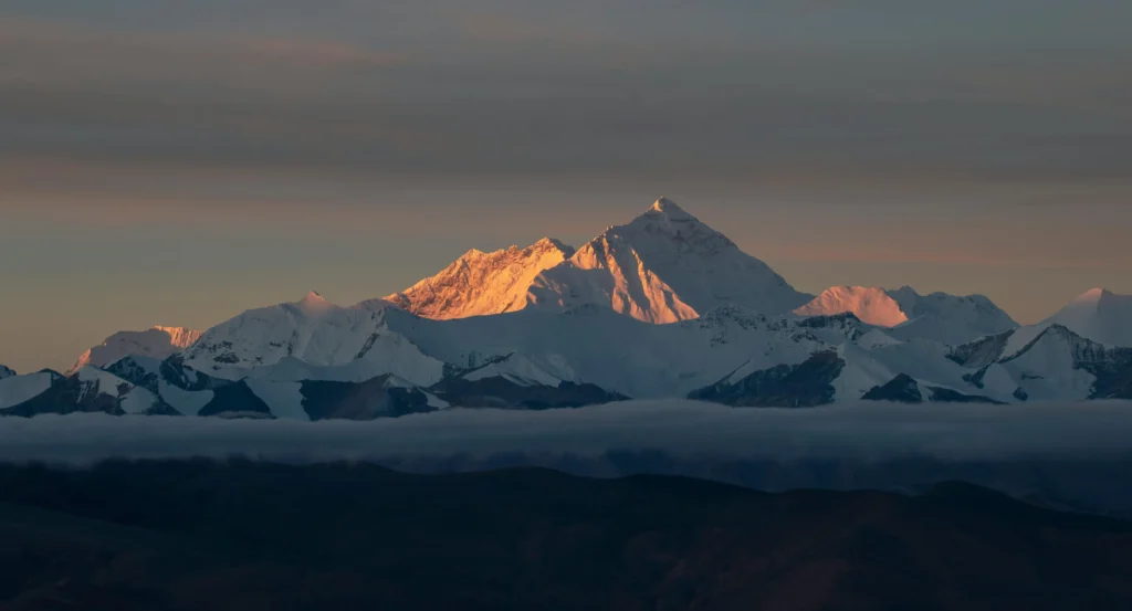 A breathtaking view of a snow-covered mountain range at sunrise or sunset, with the peaks illuminated in warm golden hues against a cloudy sky, and lower slopes shrouded in mist.