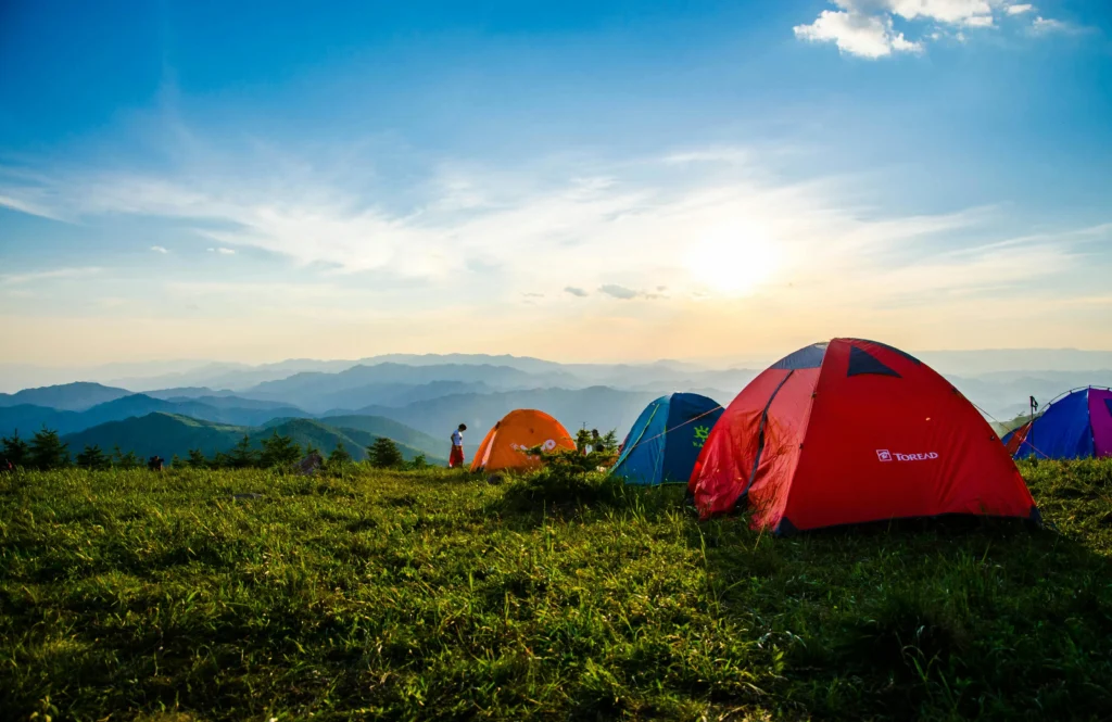 A scenic campsite at sunrise with several colorful tents pitched on a grassy hill, surrounded by rolling mountains under a clear blue sky with scattered clouds.