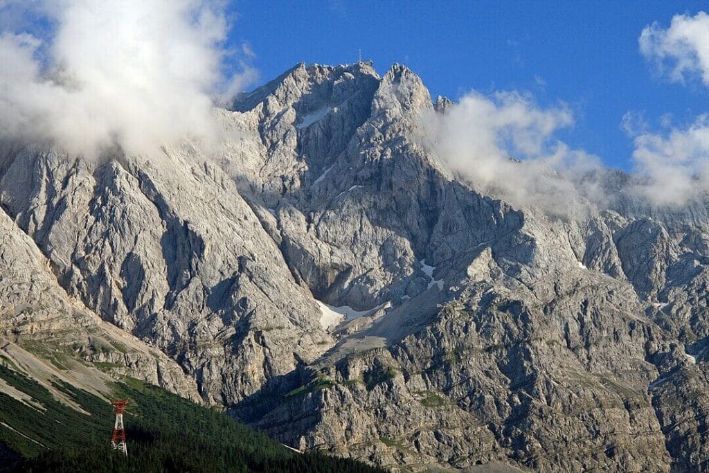 A dramatic snow-capped alpine peak towers above jagged rocky ridges under a deep blue sky. Patches of snow cling to the steep granite faces, with lower slopes revealing hints of green vegetation and distant mountain ranges fading into the horizon.