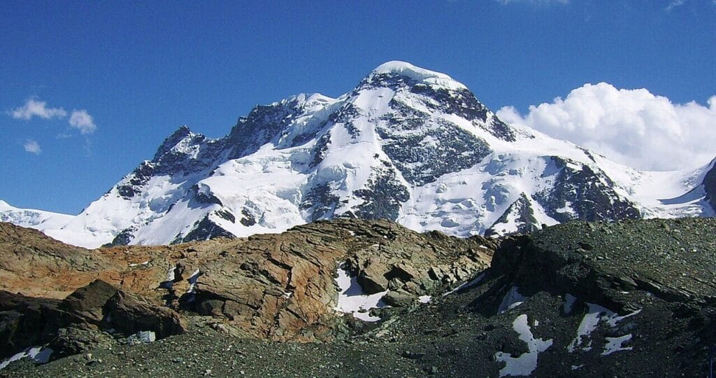 A majestic snow-capped mountain peak rises sharply against a clear blue sky, its steep, glacier-streaked faces contrasting with the rugged, rocky foreground ridge dusted in patches of snow.