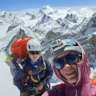 Two climbers taking a selfie on a snowy mountain ridge, with stunning views of snow-covered peaks under a clear blue sky. Both are wearing helmets, sunglasses, and climbing gear, with one carrying a large orange backpack and the other a camera attached to their helmet.