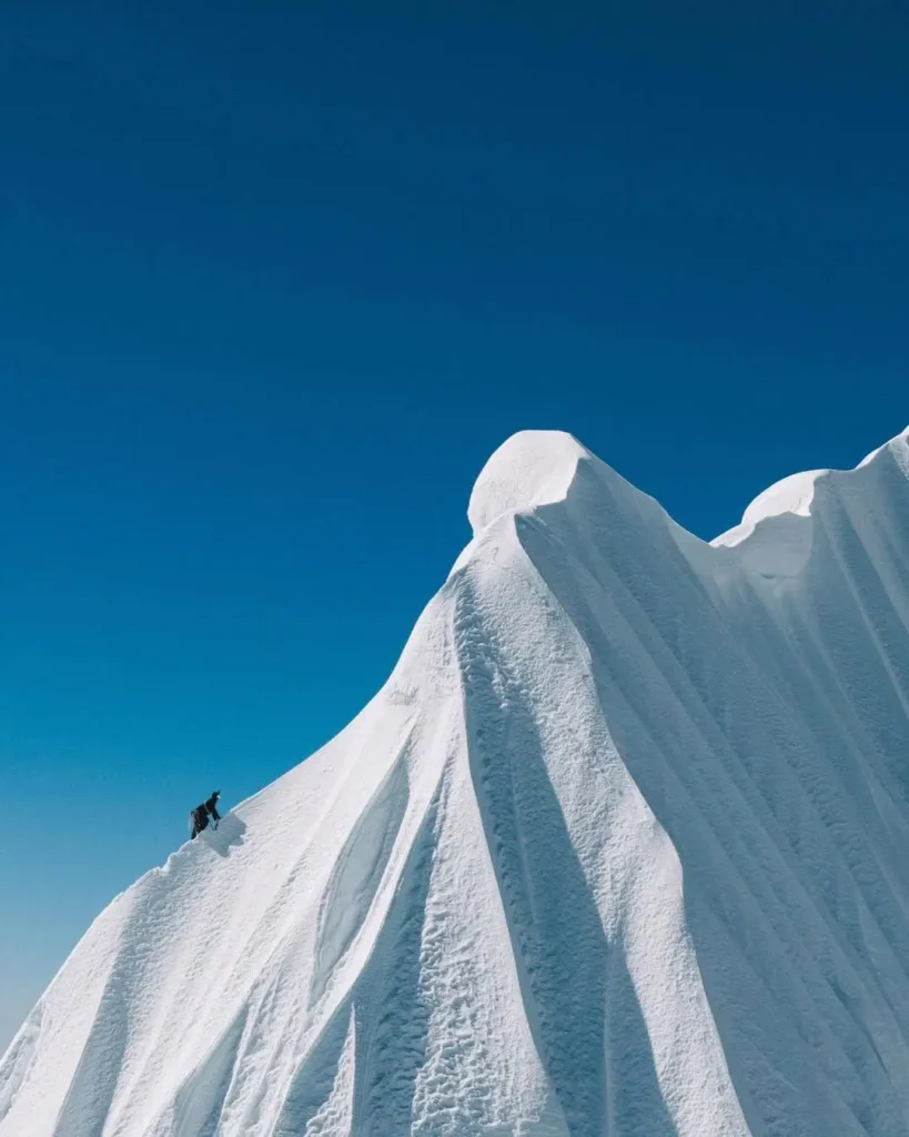 A person in dark clothing is climbing a steep, snow-covered ice formation under a clear blue sky. The ice has smooth, flowing ridges and peaks, creating a dramatic and rugged landscape.