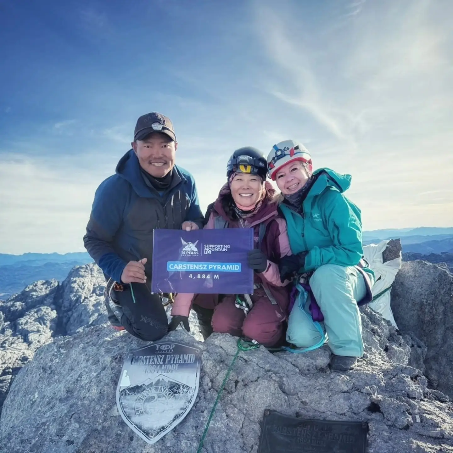 A group of three climbers, two adults and a child, posing together on the summit of Carstensz Pyramid at 4,884 meters. They are holding a sign that reads "CARSTENSZ PYRAMID 4,884 M" with the "14 Peaks" logo and "Supporting Life" text. The background features a clear sky and rugged mountain peaks.