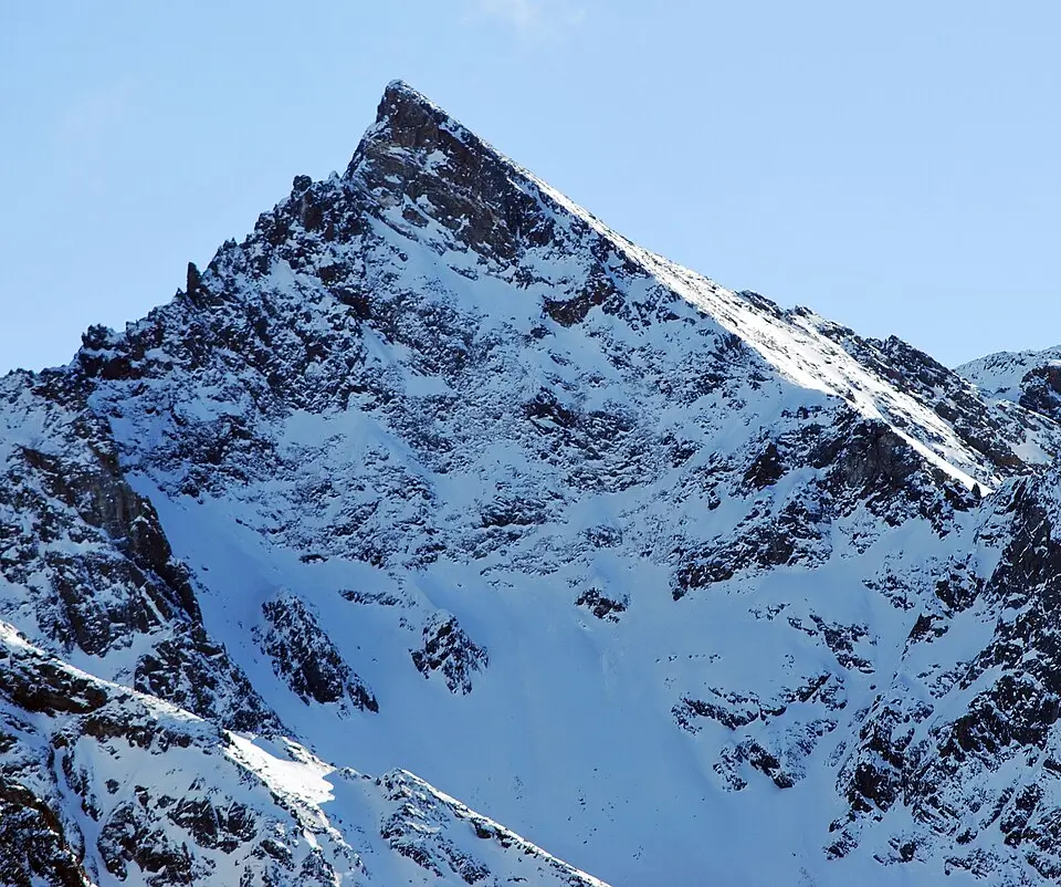 A sharply pointed, snow-covered mountain peak rises against a clear blue sky, its steep rocky slopes dusted with fresh snow and shadowed ridges creating dramatic contrast in bright sunlight.