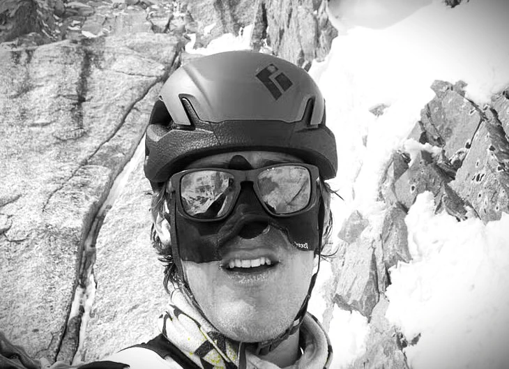 A person wearing a climbing helmet with a logo and dark sunglasses smiles at the camera, with a rugged, snow-covered rock face in the background.