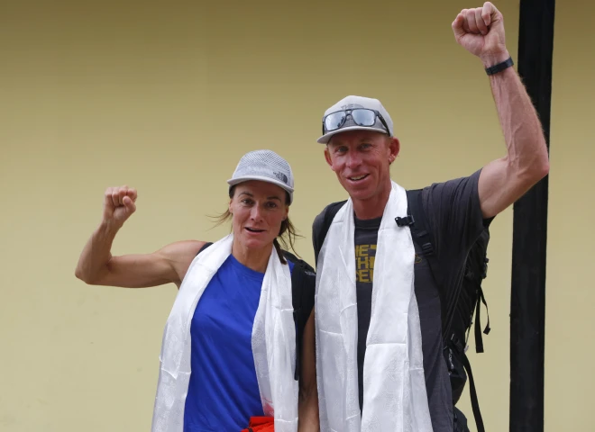 Two people standing side by side with raised fists, wearing white scarves and casual outdoor clothing, including backpacks, against a plain yellow wall.
