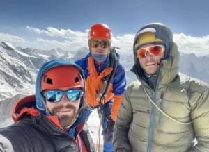 Three climbers wearing helmets and sunglasses pose for a selfie against a backdrop of snow-covered mountains under a partly cloudy sky. They are dressed in colorful mountaineering gear, including jackets and harnesses, with one holding an ice axe.