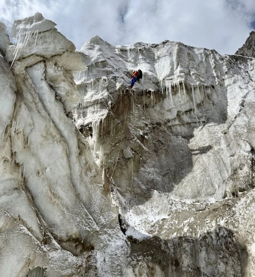a close-up of a climber in orange and blue gear ascending an icy wall with ropes, surrounded by icicles and snow.