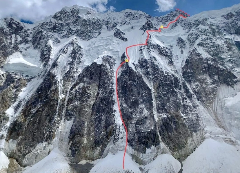 A snow-covered mountain face with a red line marking a climbing route, featuring yellow markers at key points. The route ascends steeply through icy and rocky terrain under a partly cloudy sky.