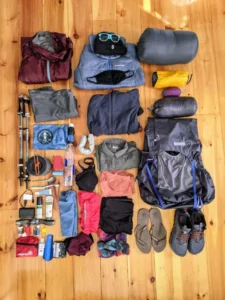 A neatly organized collection of camping and hiking gear laid out on a wooden floor. The items include a red rain jacket, a blue puffy jacket with a black face mask and sunglasses, a gray sleeping bag, a yellow tent, a purple sleeping pad, a large gray backpack, hiking poles, various clothing items (jackets, shirts, pants, underwear), a water bottle, a first aid kit, toiletries, a small pot, flip-flops, and trail running shoes.