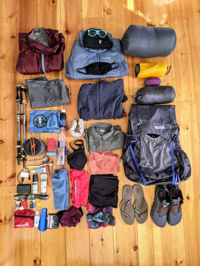 A neatly organized collection of camping and hiking gear laid out on a wooden floor. The items include a red rain jacket, a blue puffy jacket with a black face mask and sunglasses, a gray sleeping bag, a yellow tent, a purple sleeping pad, a large gray backpack, hiking poles, various clothing items (jackets, shirts, pants, underwear), a water bottle, a first aid kit, toiletries, a small pot, flip-flops, and trail running shoes.