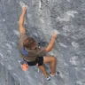 A person rock climbing on a steep, rugged cliff face, using both hands and feet to grip the rock. They are wearing a brown shirt, black shorts, and climbing shoes, with a harness and gear attached, including an orange chalk bag. A rope is secured to the harness and anchored to the rock above.