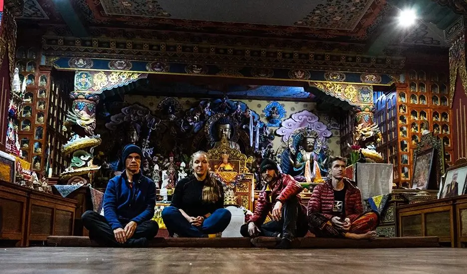 Four people sitting cross-legged on the floor of an ornate temple, surrounded by intricate decorations, statues, and colorful artwork, with a large central altar adorned with religious figures.