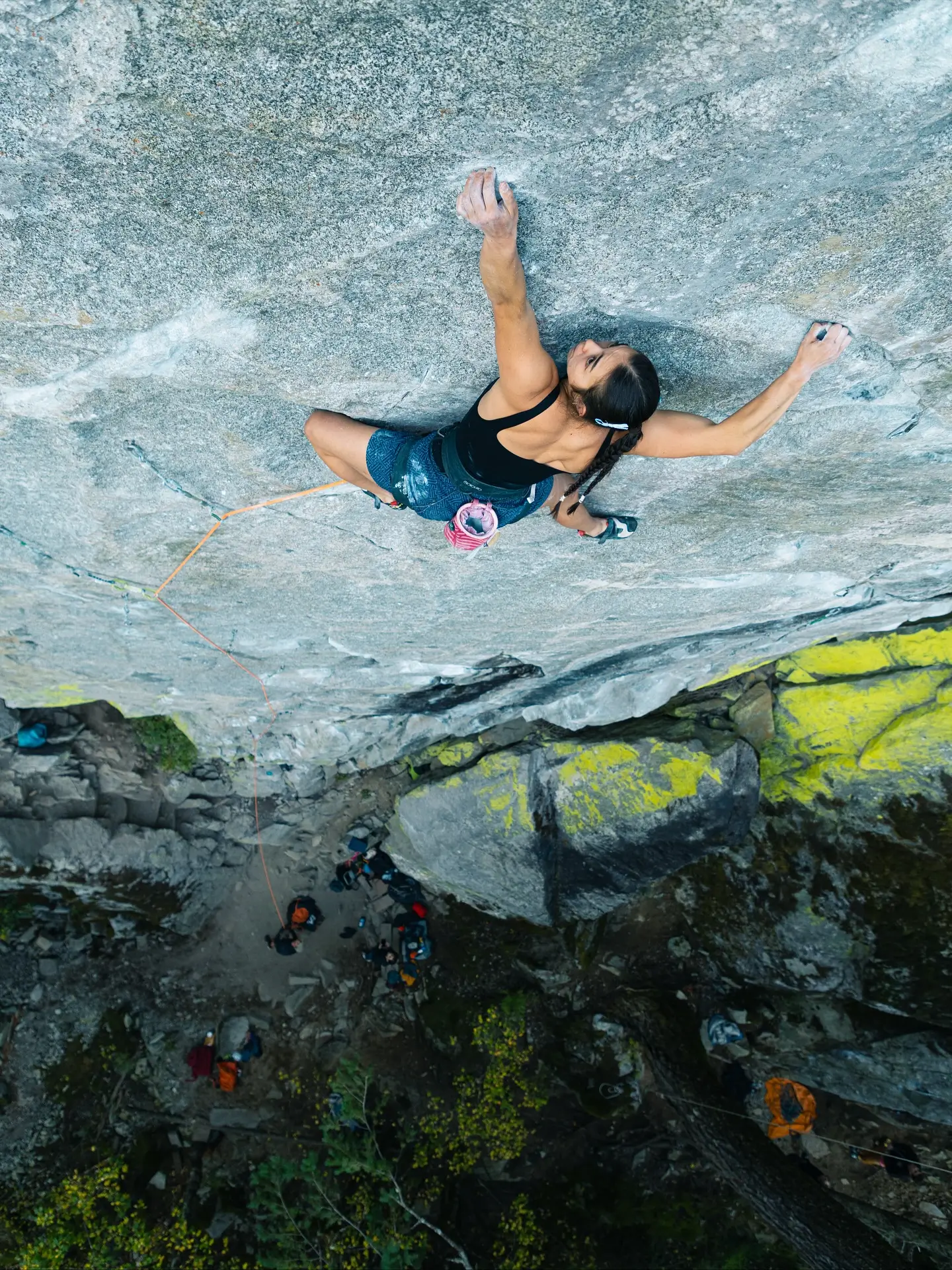 A rock climber ascends a steep granite cliff, gripping the rock face with both hands and feet. The climber is secured with an orange rope and wears a black top, blue shorts, and a chalk bag. Below, a group of people and camping gear are visible at the base of the cliff, surrounded by green vegetation and yellow lichen on the rocks. The scene is captured from an overhead perspective, highlighting the height and rugged terrain.