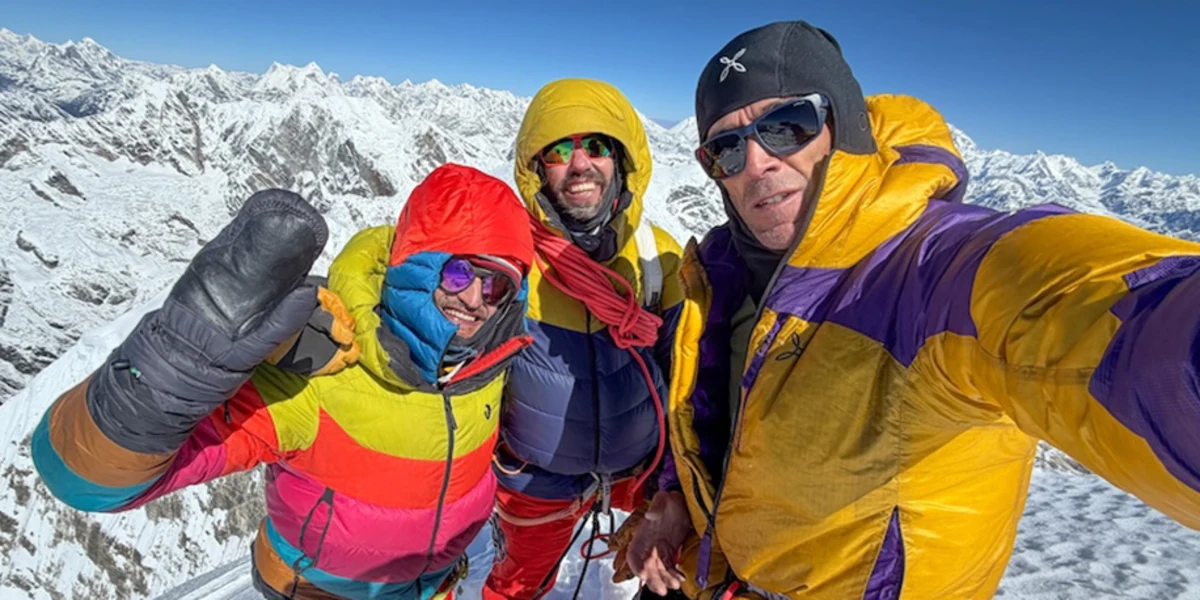 Three climbers in colorful mountaineering gear pose together on a snowy peak, with a stunning view of snow-covered mountains and a clear blue sky in the background.