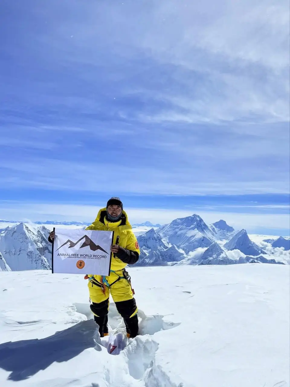 A person in a bright yellow mountaineering suit stands on a snowy mountain peak, holding a flag that reads "ANIMAL-FREE WORLD RECORD" with a mountain graphic and additional text. The background features a stunning view of snow-covered mountain ranges under a clear blue sky with some clouds.