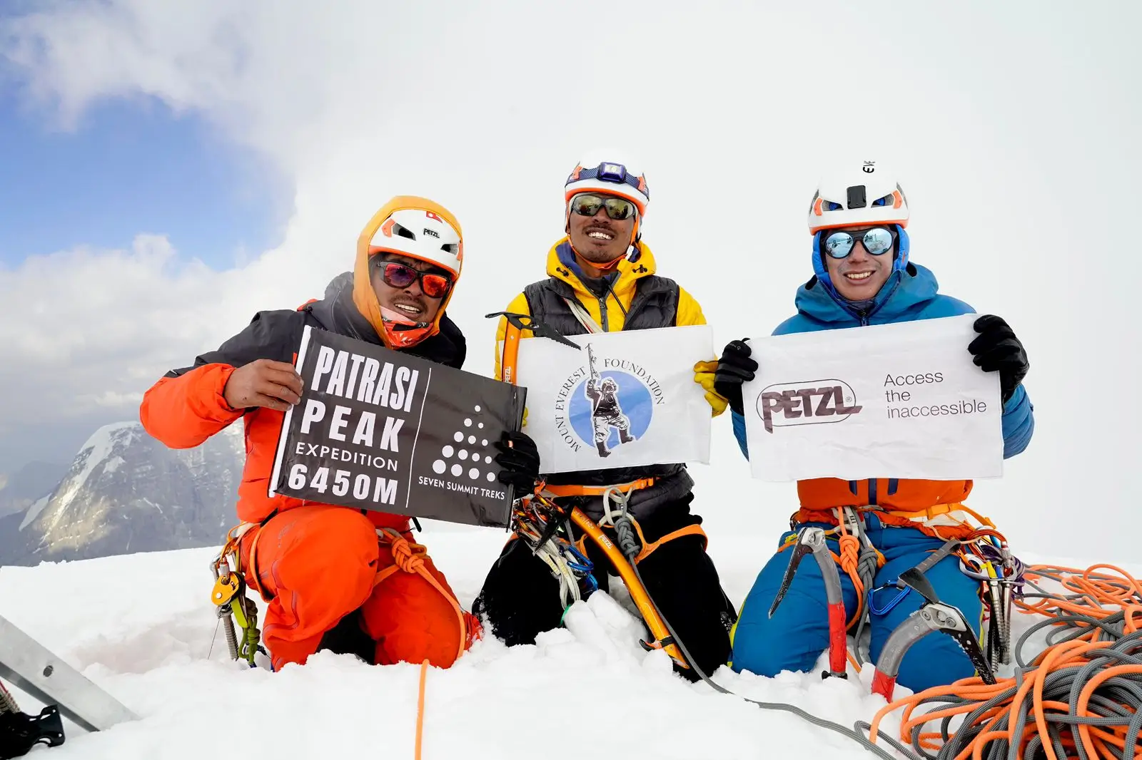 Three climbers in colorful gear sit on a snowy mountain peak, holding signs. The left climber in an orange suit holds a sign reading "Patrasi Peak Expedition 6450M." The middle climber in a yellow and black jacket holds a sign with the Mountain Foundation Seven Summits Treks logo. The right climber in a blue suit holds a sign reading "Access the inaccessible Petzl." Snow-covered mountains and a clear sky are in the background.