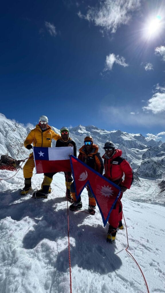 A group of four climbers stands on a snowy mountain ridge, holding flags of Chile and Nepal. They are dressed in colorful mountaineering gear, including helmets and jackets, with a bright blue sky and snow-covered peaks in the background. The sun shines brightly above scattered clouds.