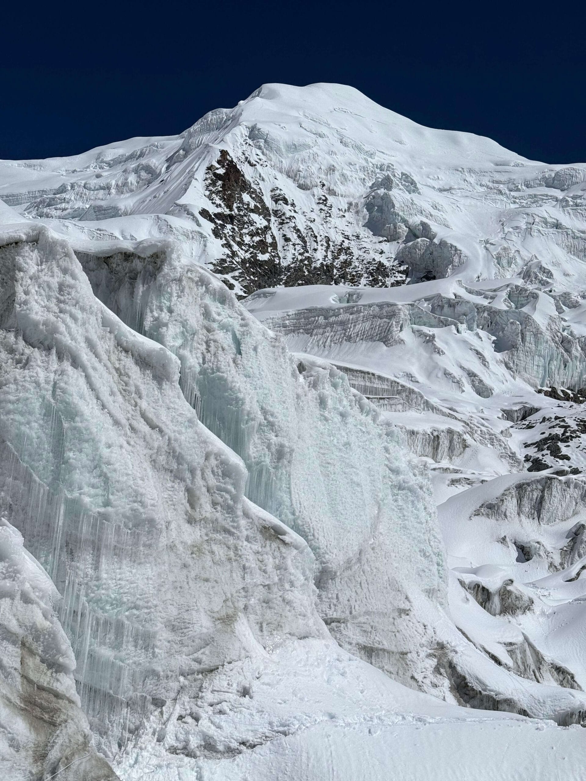 A close-up view of a rugged, snow-covered mountain landscape featuring steep ice formations and glaciers. The scene is dominated by white snow and ice against a deep blue sky, with rocky outcrops visible beneath the snow.
