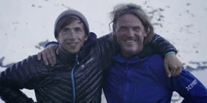 Two climbers in winter gear stand on a snowy mountain, smiling with their arms around each other. The climber on the left wears a dark jacket with blue accents and a beanie, while the climber on the right wears a blue jacket with "Eiger Extreme" branding. A snowy landscape stretches out in the background.