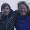 Two climbers in winter gear stand on a snowy mountain, smiling with their arms around each other. The climber on the left wears a dark jacket with blue accents and a beanie, while the climber on the right wears a blue jacket with "Eiger Extreme" branding. A snowy landscape stretches out in the background.