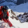 A climber in a red jacket and blue helmet ascends a steep, snow-covered mountain slope, secured with an orange rope. The background features rugged, snow-dusted peaks under a clear sky.