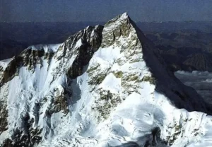 A majestic snow-covered mountain peak rising sharply against a clear blue sky, with rugged slopes and ridges, and a vast expanse of lower mountain ranges and clouds visible in the background.